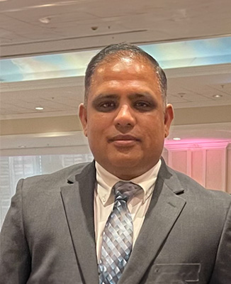 The image shows a man standing and smiling at the camera. He is wearing a suit, with a tie visible, and appears to be at an event or conference given the background that suggests an indoor setting with a stage and lighting rigs.