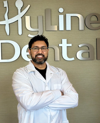 The image shows a man in a white lab coat standing confidently with his arms crossed, smiling at the camera, in front of a sign that reads  HYLINE DENTAL.