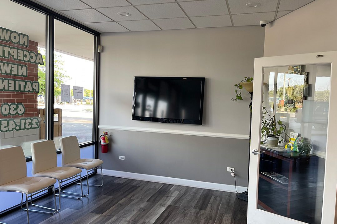 The image shows an interior view of a modern waiting area with a flat-screen TV mounted on the wall, a seating arrangement consisting of chairs and a sofa, a reception desk, and a glass display case.