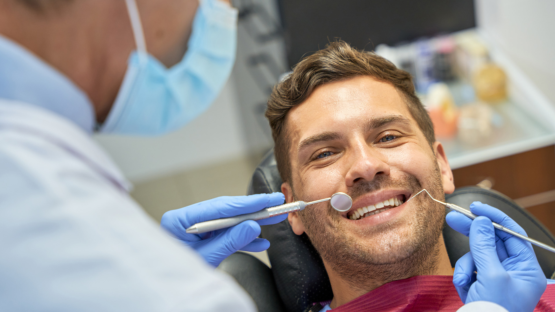 The image features a man sitting in a dental chair, receiving dental treatment. He is smiling and appears to be relaxed during the procedure. A dentist is actively engaged in cleaning his teeth while wearing a face mask and protective gloves.