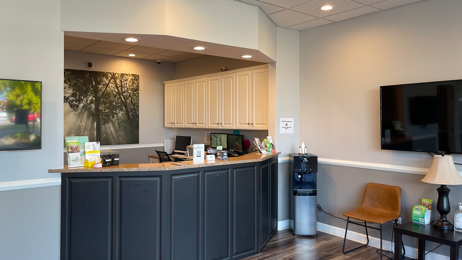 The image depicts an interior view of a well-lit office space with a modern design, featuring a reception area with a counter, chairs, and a desk, as well as a waiting area with a television screen displaying a landscape image.