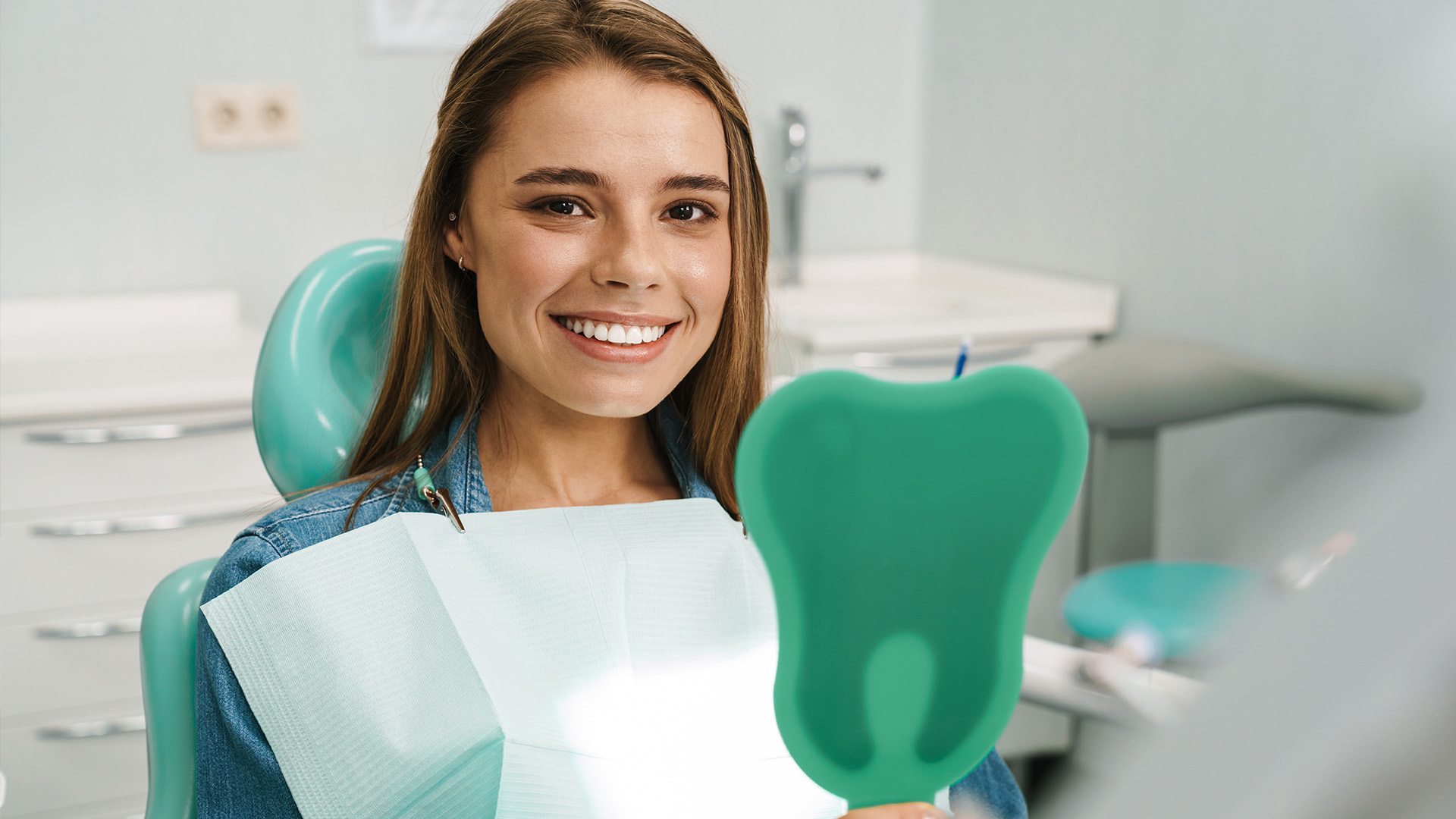 A young woman in a dental office, smiling and holding a toothbrush-shaped object, wearing a blue shirt.