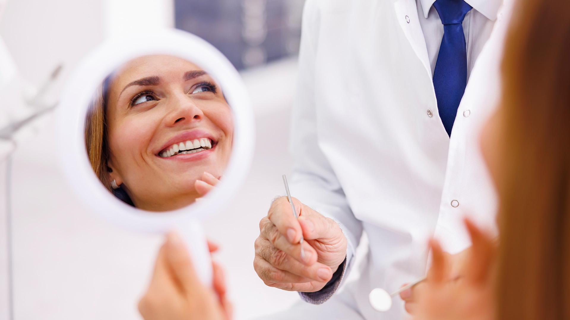 A dentist is smiling at a woman in a dental chair, both looking into a large mirror.