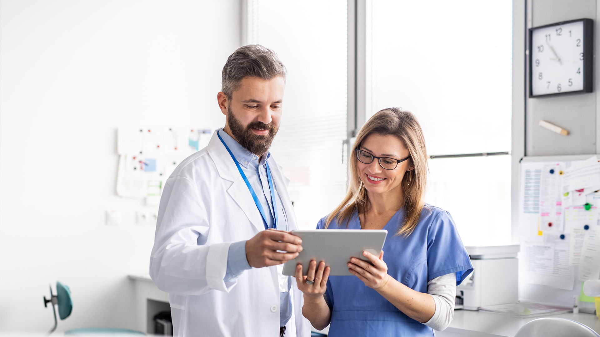 A male and female healthcare professional, likely a doctor and nurse, in a hospital setting, reviewing digital information on a tablet.