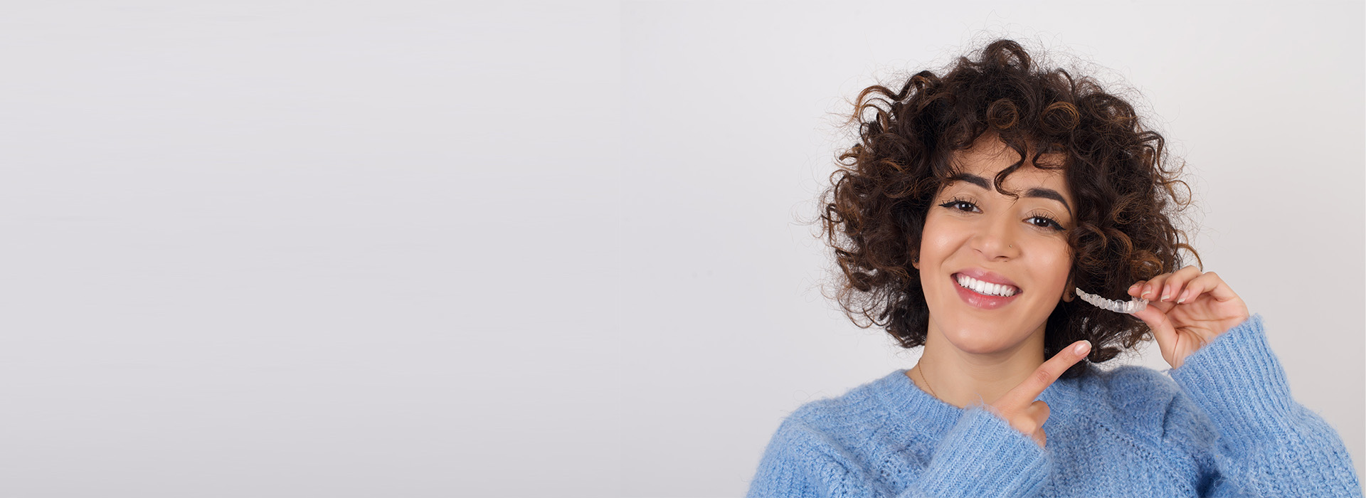 The image is a photograph of a smiling woman with curly hair, holding a smartphone to her ear, against a plain background.