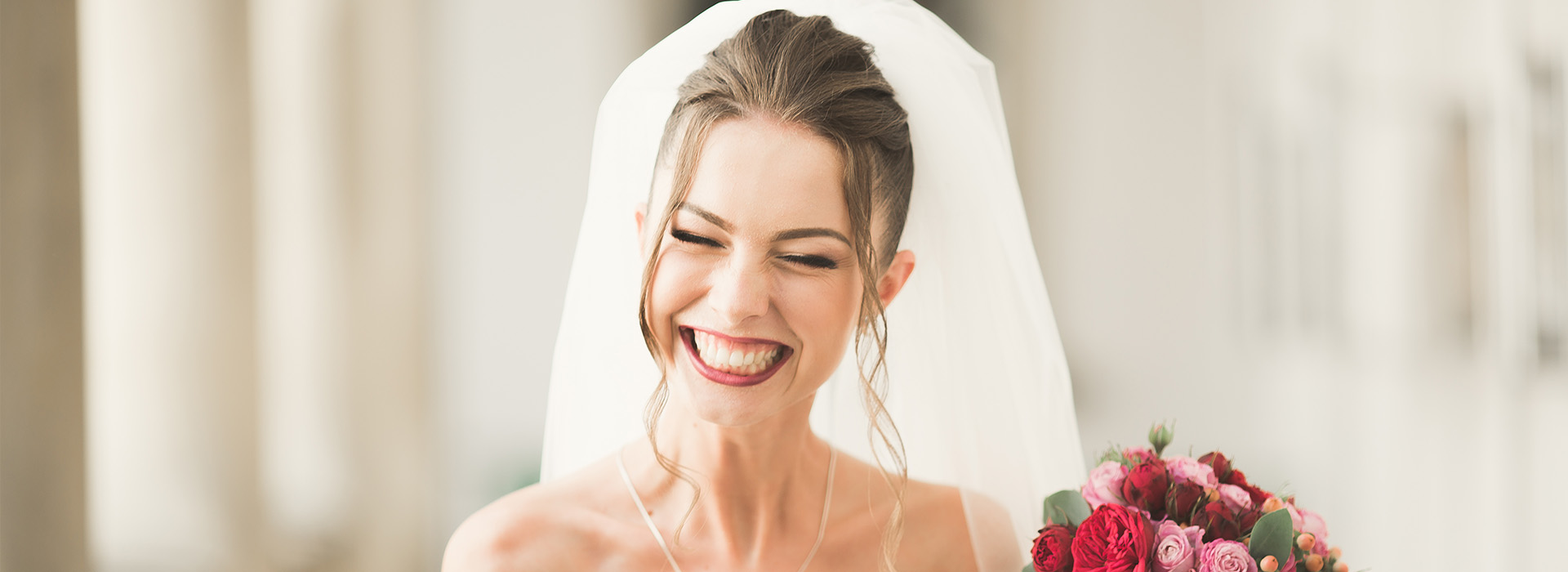 The image is a photograph of a woman wearing a wedding veil and smiling, standing in front of a blurred background that suggests an indoor setting.