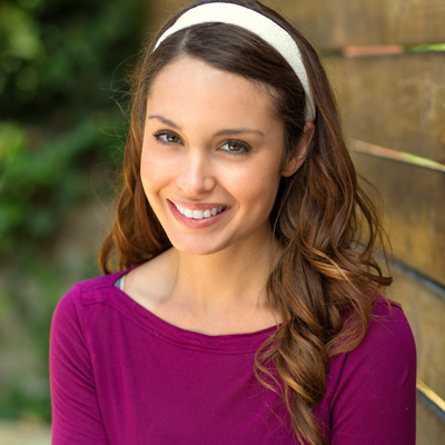 Smiling woman with long hair, wearing a headband and pink top, posing for portrait against wooden fence backdrop.