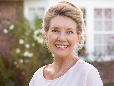 The image is a portrait of an older woman with light hair, wearing a white top and smiling at the camera. She has a fair complexion and is standing outdoors in front of a house with a visible brick facade.