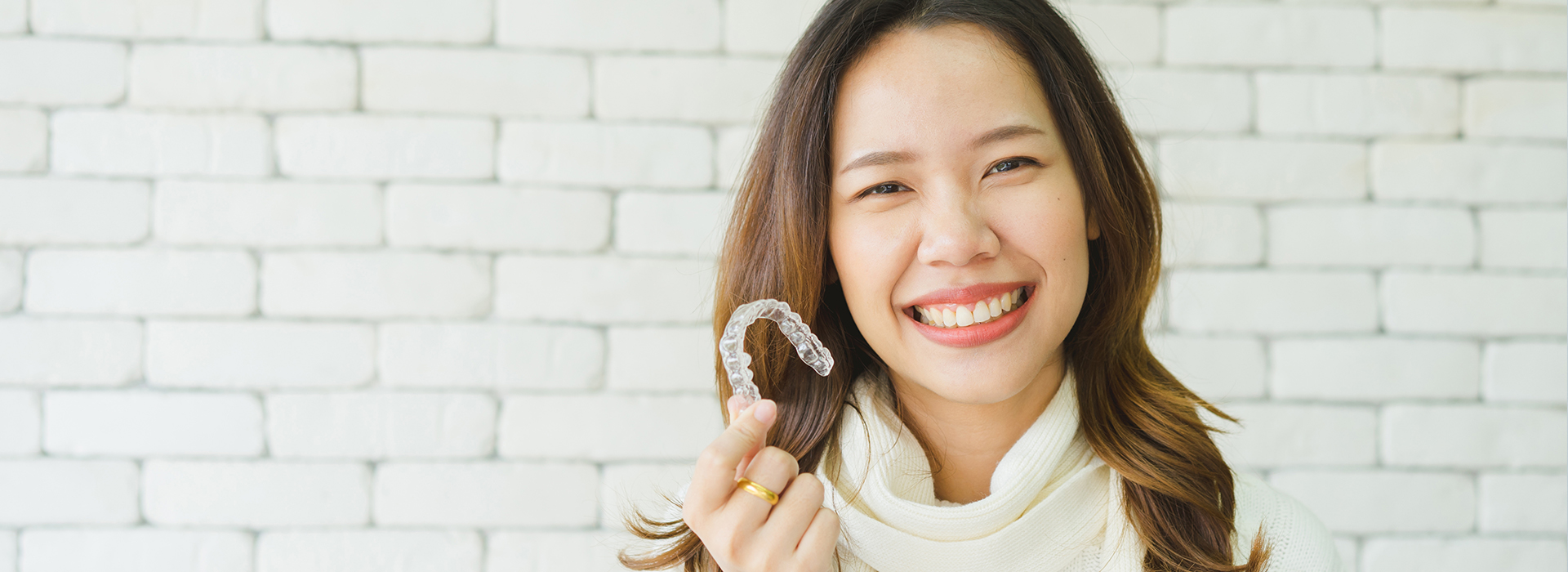 A woman with a smile is holding a ring close to her face, set against a white brick wall.