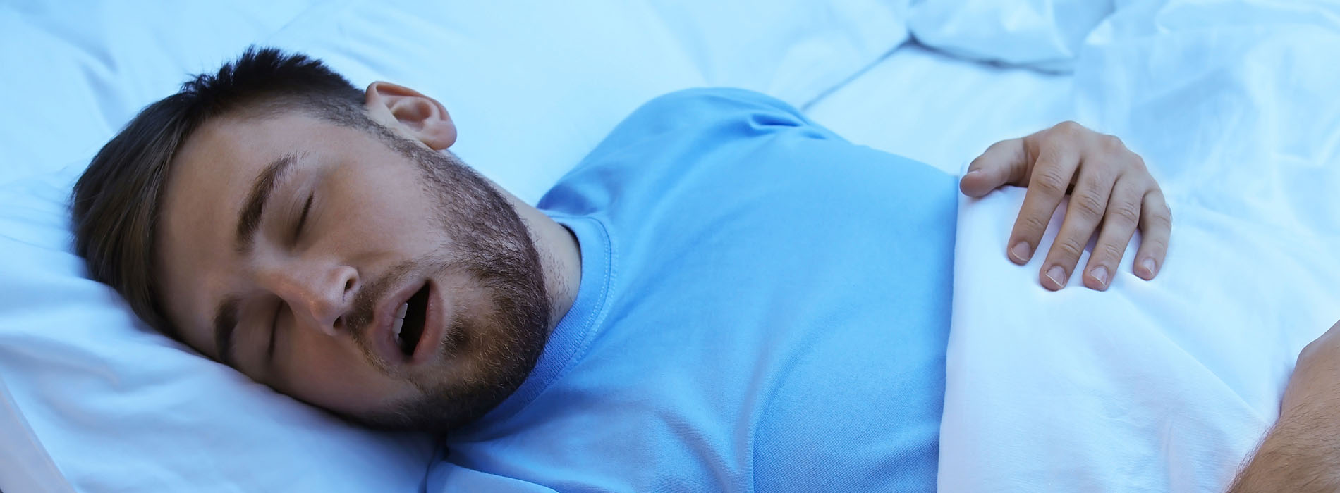 A man in a blue shirt sleeping on his side with his head turned towards the camera, lying in bed against a white background.