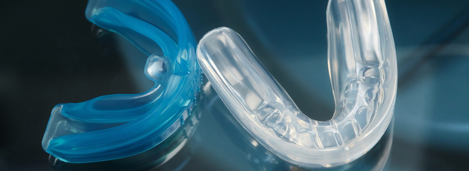 The image shows a clear, blue-tinted acrylic dental retainer with a design that resembles the letter  S  and is placed against a blurred background.