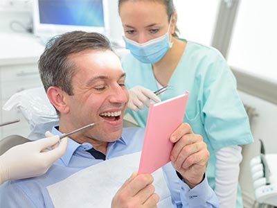 A man in a dental chair, holding up a pink card with a smile, while a dentist is looking at him and another person is taking a photo.