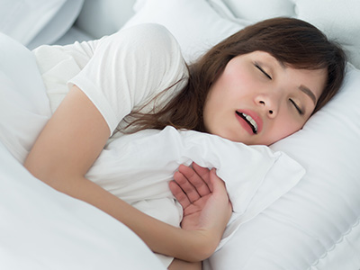 A woman in a white shirt is lying down in bed, her eyes are closed and she appears to be asleep with her mouth slightly open.